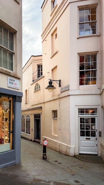A narrow street scene shows the exterior of a small, multi-storey property in Mayfair with cream-colored walls and several large, multi-pane windows. Adjacent to the building, a shopfront with a large glass window and a sign reading 'Avery Howe W1' is visible on the left. The scene is captured during daylight with natural light illuminating the street and buildings. A black outdoor wall lantern extends from the corner of the building, and a small street sign indicating the street name is mounted above a white door with a window at the ground level. The pavement is paved with small stone tiles, and a black and yellow bollard is positioned in the middle of the street to prevent vehicle entry. This setting illustrates the typical environment for home relocation or furniture transport, with no visible moving equipment or personnel present. The image represents the urban context suitable for small flat moves in Mayfair, undertaken by removal specialists such as Mayfair Removals, focusing on efficient space management and transport logistics for city-based house removals.
