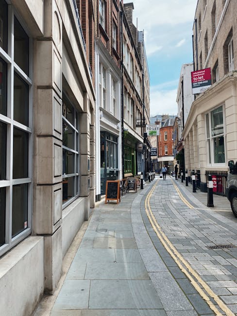 A narrow street scene shows the exterior of a small, multi-storey property in Mayfair with cream-colored walls and several large, multi-pane windows. Adjacent to the building, a shopfront with a large glass window and a sign reading 'Avery Howe W1' is visible on the left. The scene is captured during daylight with natural light illuminating the street and buildings. A black outdoor wall lantern extends from the corner of the building, and a small street sign indicating the street name is mounted above a white door with a window at the ground level. The pavement is paved with small stone tiles, and a black and yellow bollard is positioned in the middle of the street to prevent vehicle entry. This setting illustrates the typical environment for home relocation or furniture transport, with no visible moving equipment or personnel present. The image represents the urban context suitable for small flat moves in Mayfair, undertaken by removal specialists such as Mayfair Removals, focusing on efficient space management and transport logistics for city-based house removals.